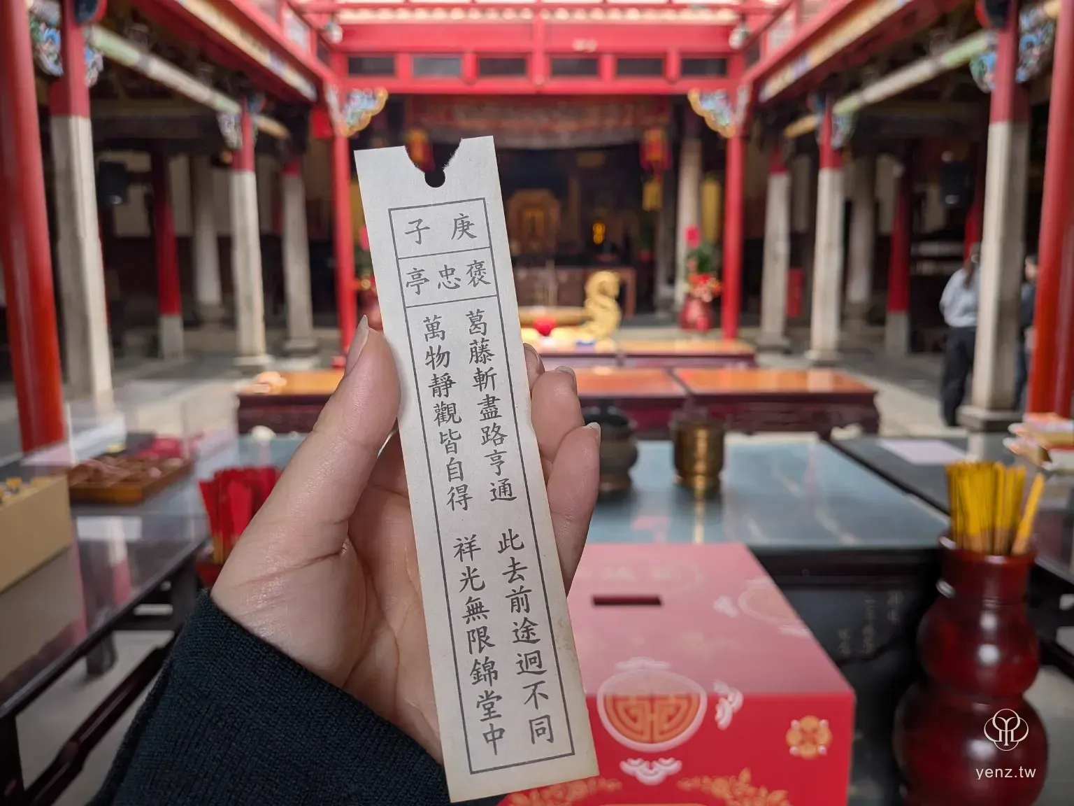 手中拿著義民廟第 37 籤籤詩，背景是模糊的廟內供桌與紅色柱列。