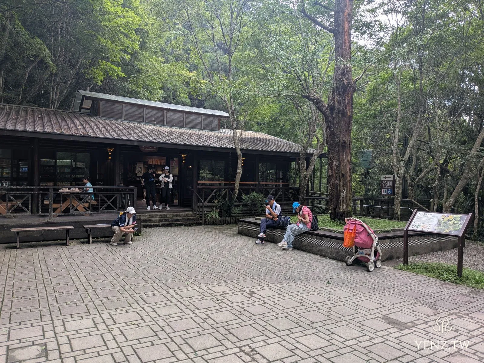 森林小舖外觀，木構建築與鋪面廣場，有遊客坐在長凳歇腳。