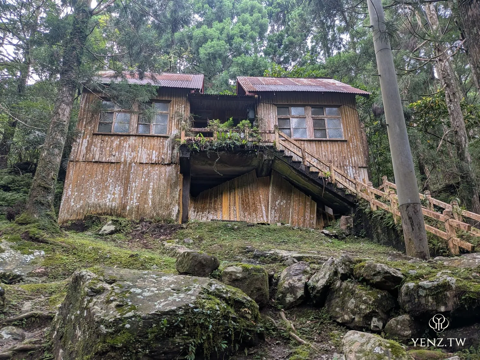 林中一棟廢棄的兩層木屋，鏽蝕的鐵皮屋頂，旁邊有木造階梯。