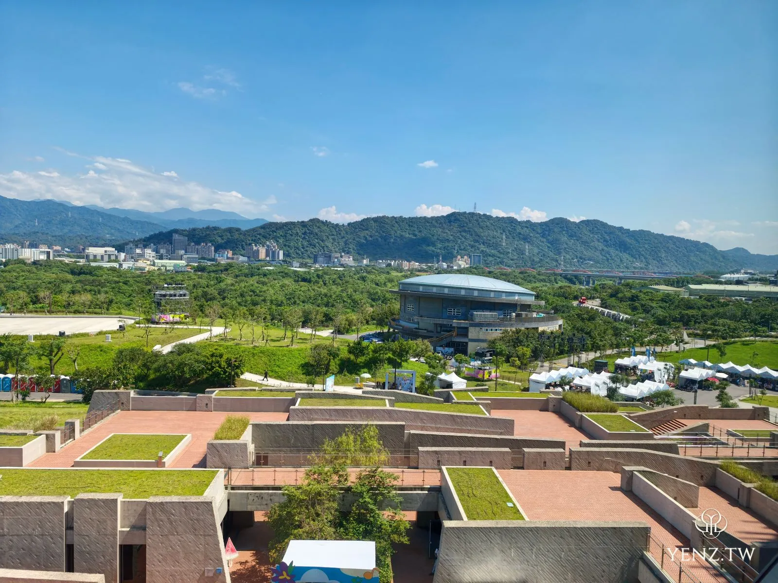 從高處俯瞰整片園區，紅磚清水模的地景屋頂、綠草坪、白色市集帳篷群與遠山。