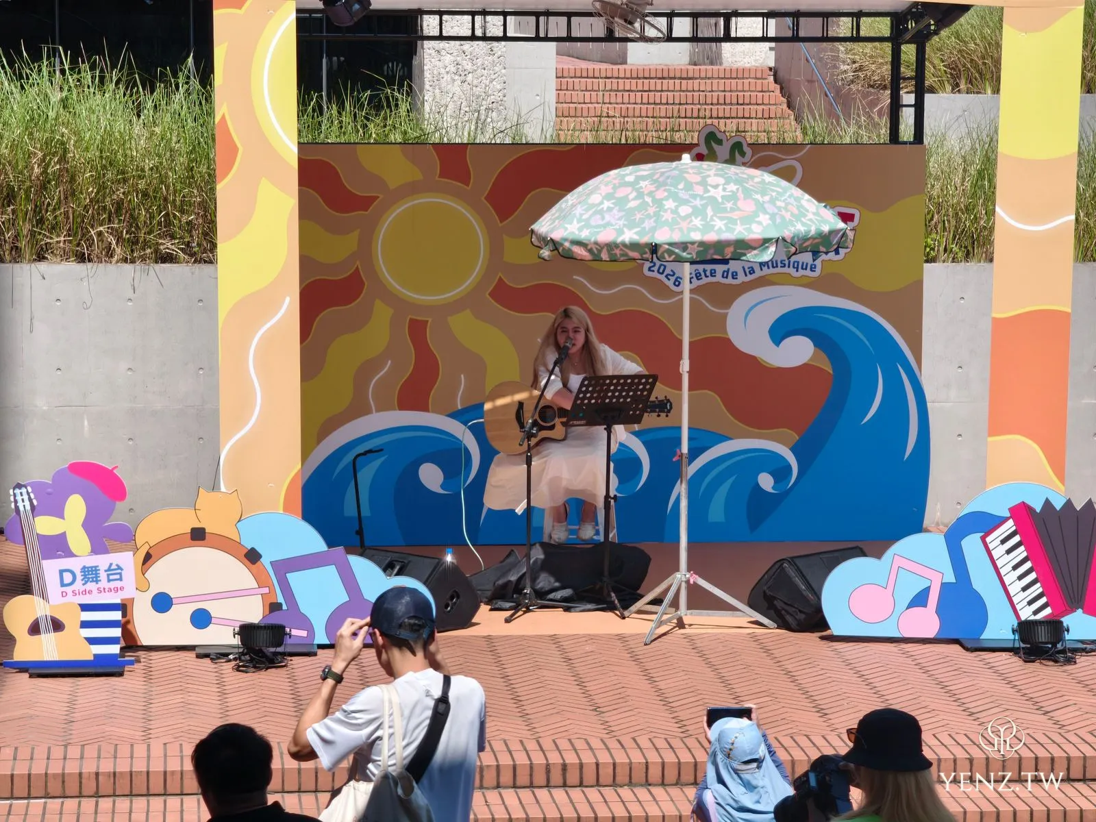 D 舞台上一位女歌手抱著木吉他演出，背板畫著太陽與海浪。