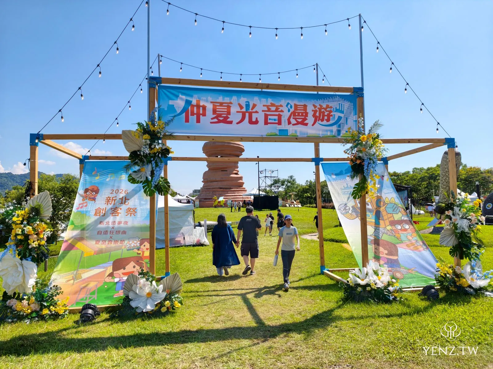 草地上一座木頭拱門寫著「仲夏光音漫遊」，下方橫幅是「2026 新北創客祭」，背後有堆疊的陶藝圓雕。